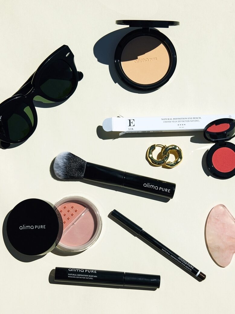 A flat lay of assorted makeup items, including powder, blush, lipstick, eyeliner, a brush, sunglasses, a gua sha tool, and earrings, arranged on a light background.