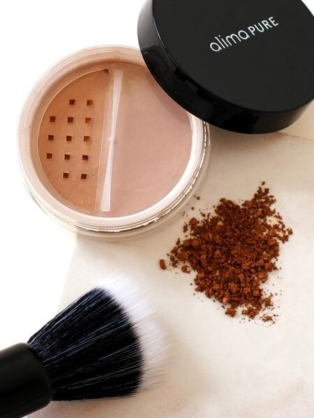 Loose powder container with divided sections, scattered powder, and a makeup brush on a light surface.