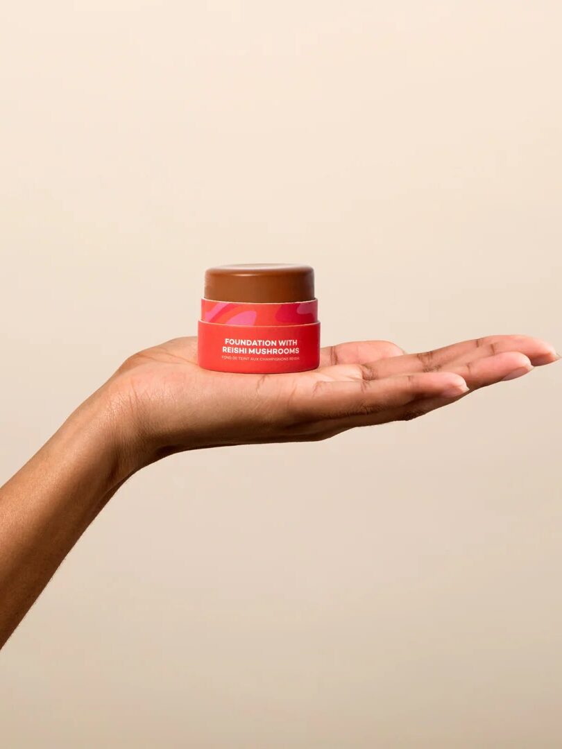 A hand holds a small container of foundation with reishi mushrooms against a beige background.