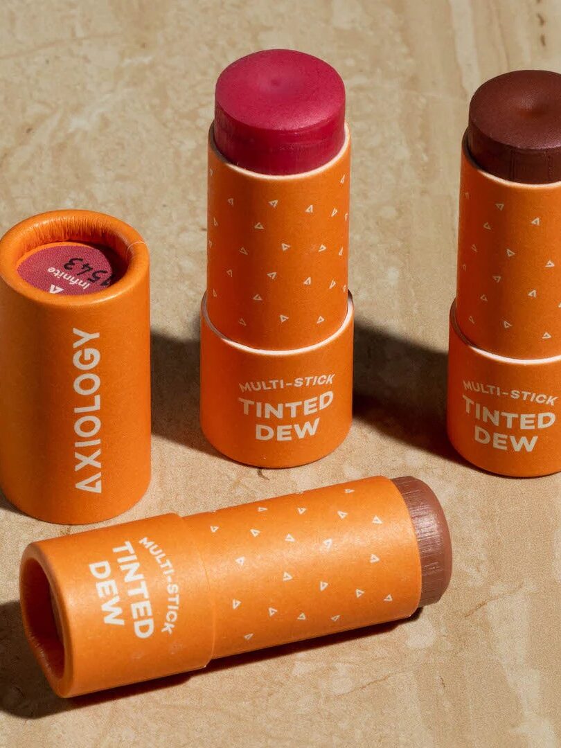 Three orange tubes of Axiology Tinted Dew Multi-Stick in various shades lie on a wooden surface.