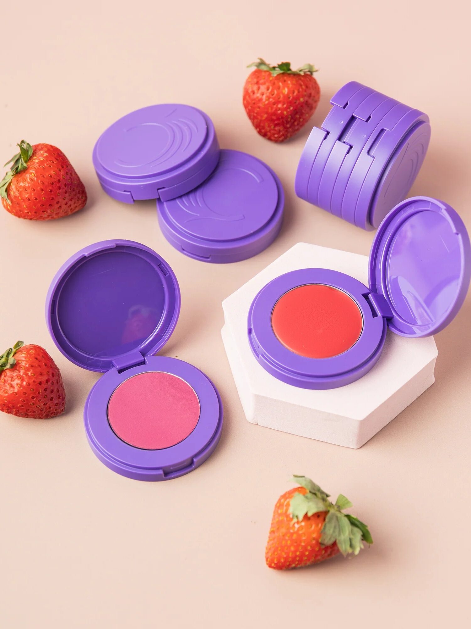 Purple cosmetic compacts with blush and strawberries on a pink background.