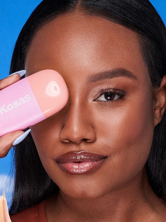 Person holding a pink Kosas product near their face against a blue background with a swatch of product shown at the bottom.