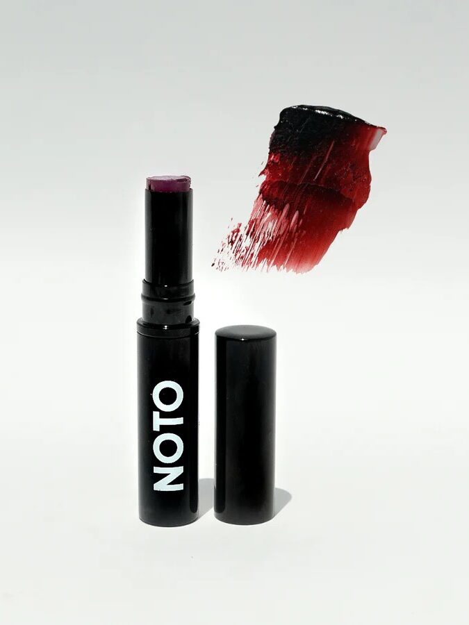 Black lipstick tube labeled "NOTO" with cap off and a swipe of dark red lipstick above it against a white background.