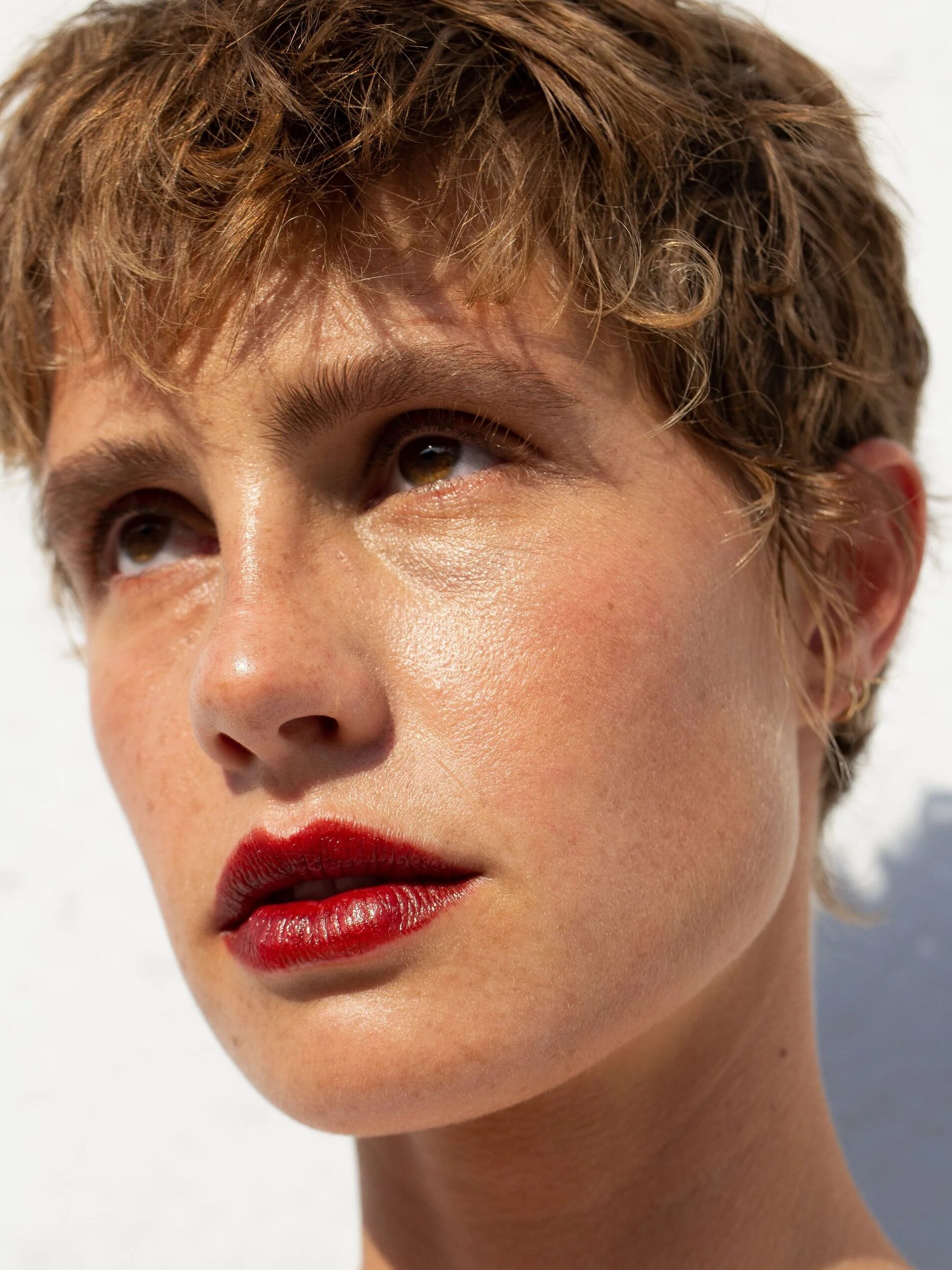 A person with short, tousled hair and red lipstick looks into the distance against a bright background.