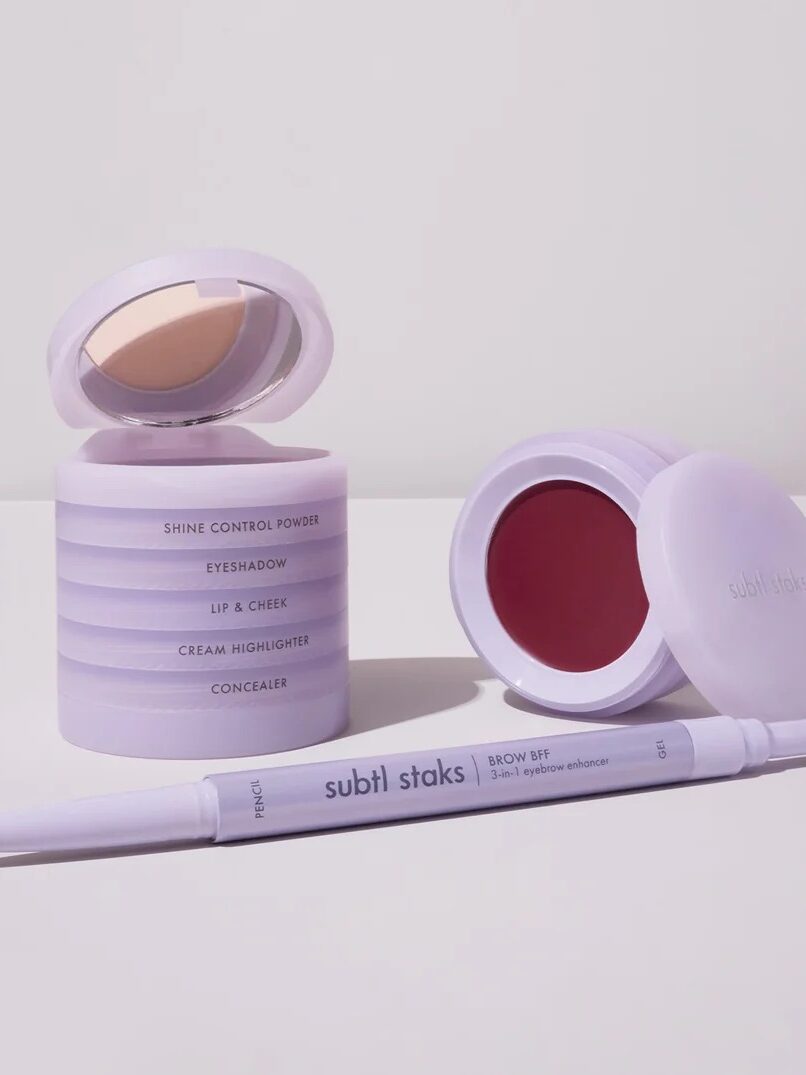 A set of cosmetic products with a stack of round compacts, one open showing a mirror, and a dual-ended pencil with a brush, all in light purple packaging.