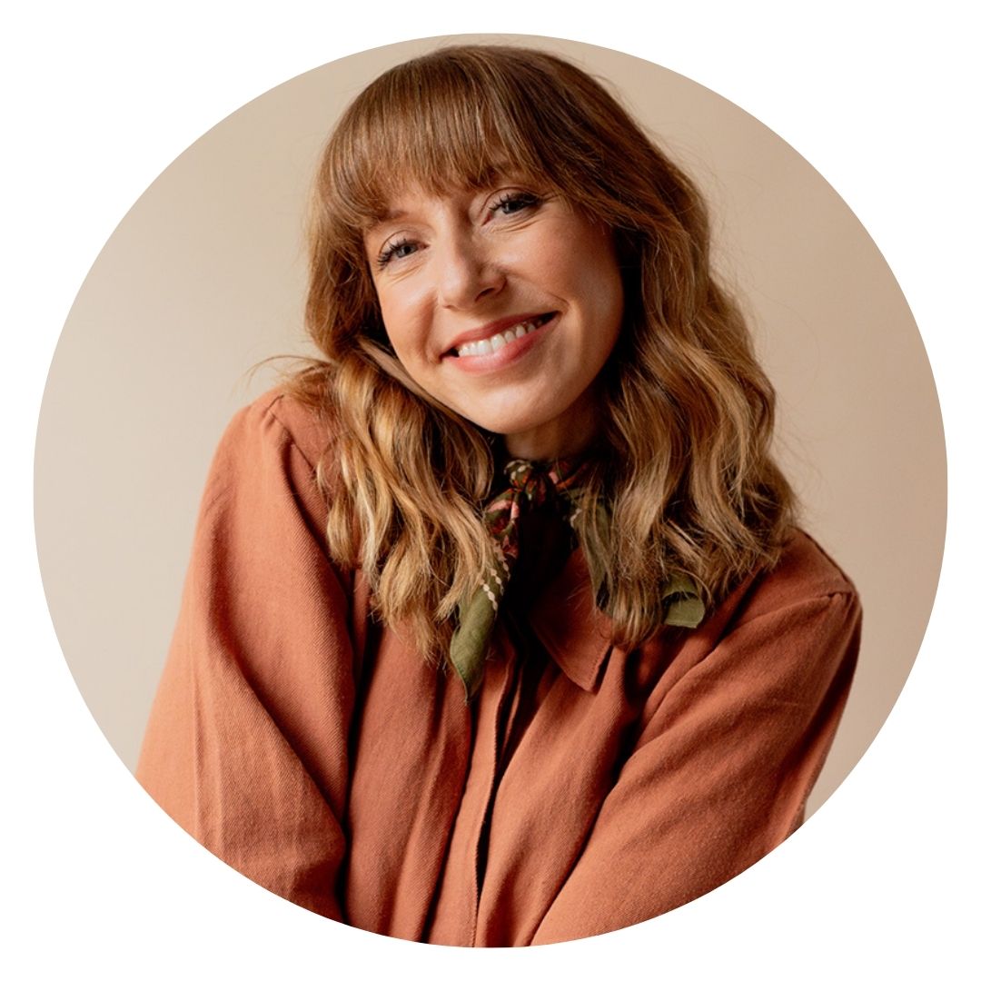 Courtney Jay Higgins has wavy, shoulder-length hair and wears a rust-colored top and a green scarf, smiling at the camera.