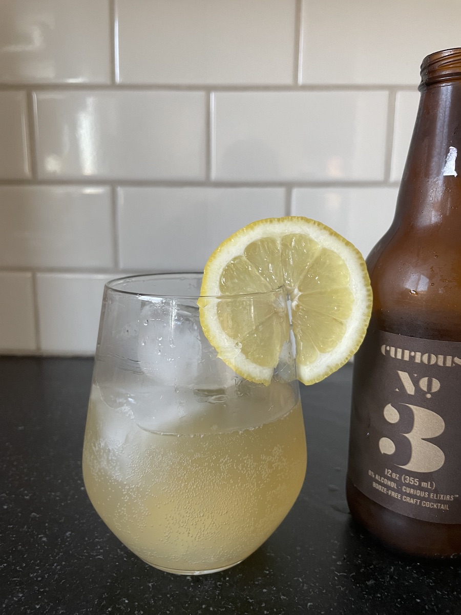 A glass of chilled beverage with ice and a lemon slice on the rim, next to a brown bottle, set against a tiled backsplash.