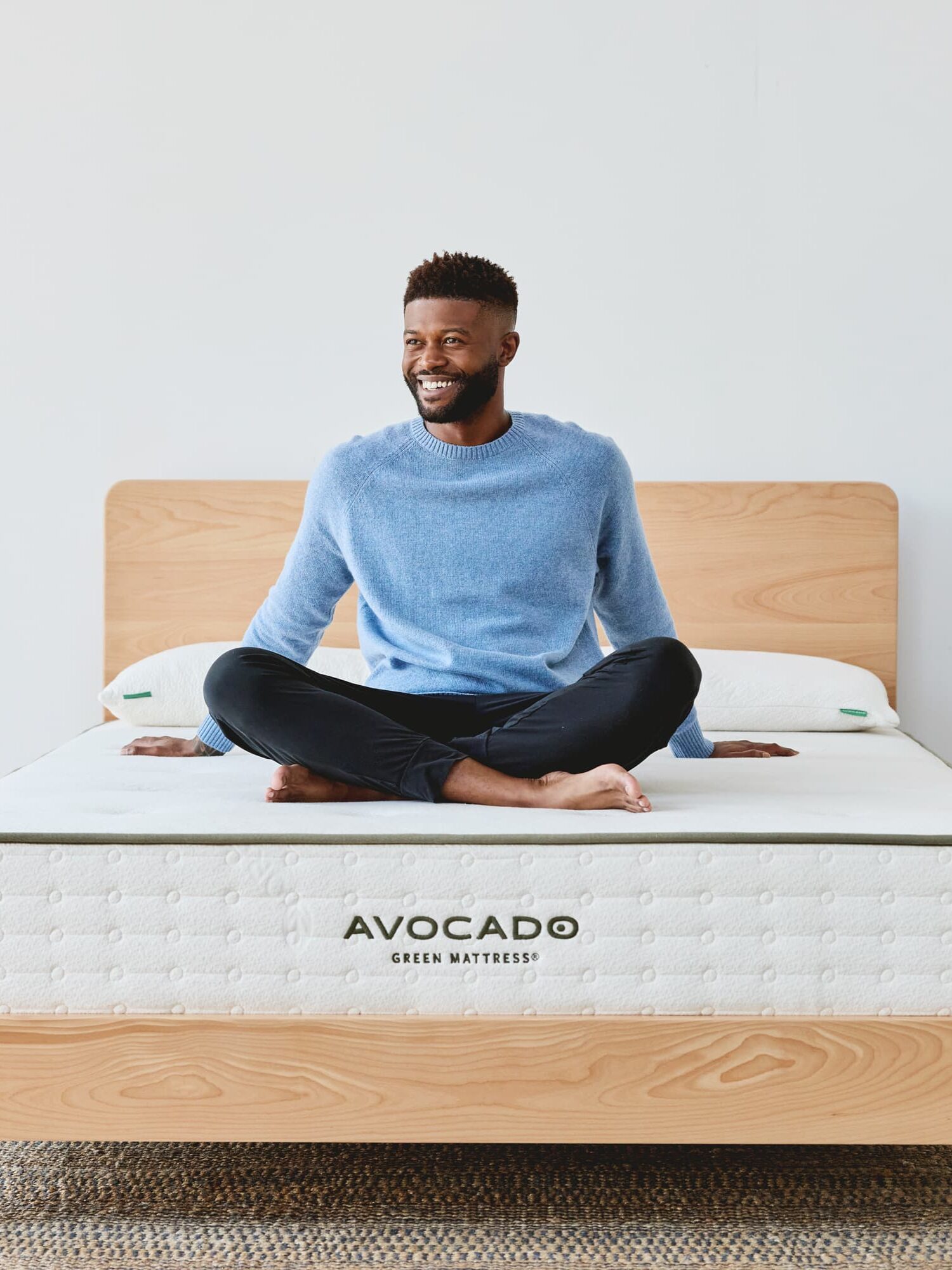 Man in a blue sweater sits cross-legged on an Avocado Green Mattress atop a wooden bed frame in a bright room.