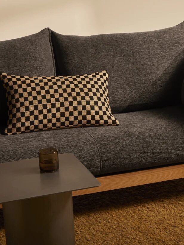 A gray sofa with a checkered cushion next to a small, square table with a glass on it, set on a textured mustard-yellow rug.