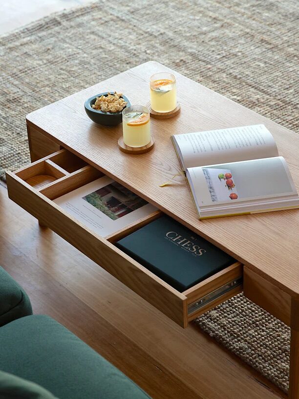 Wooden coffee table with open drawer containing books. Two candles and a bowl of snacks on top. Open book displayed. Nearby green sofa on a textured rug.