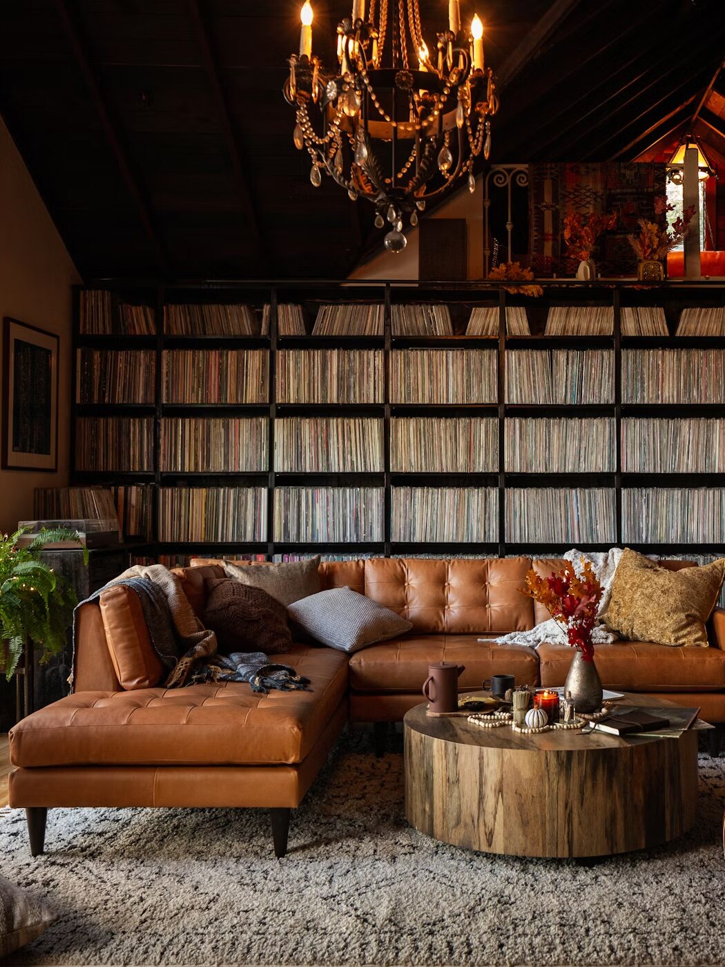 A cozy living room with a brown leather sofa, circular wooden coffee table, and shelves filled with records. A chandelier hangs from the ceiling, and a drum set is visible on the right.