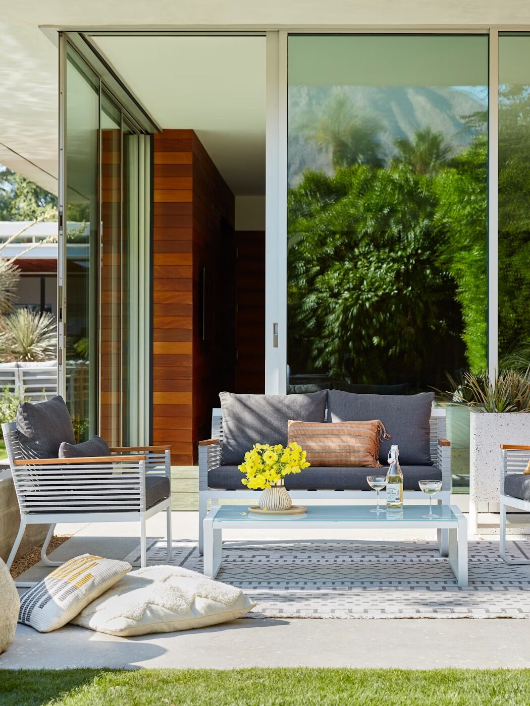 Modern patio with a glass door, wooden panels, and white metal furniture. A table with flowers and drinks is set on a patterned rug. Surrounding greenery and cushions add to the cozy atmosphere.