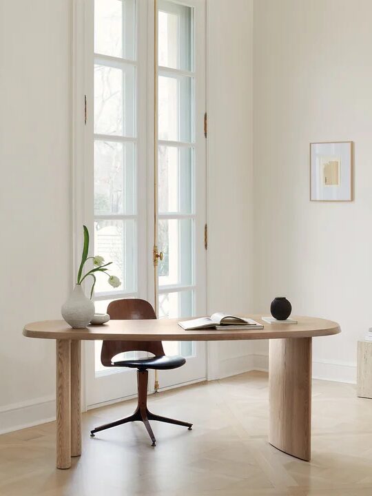 A minimalist home office features a wooden desk with a chair, a vase with a plant, an open book, and a small speaker. A large window and a framed picture are in the background.