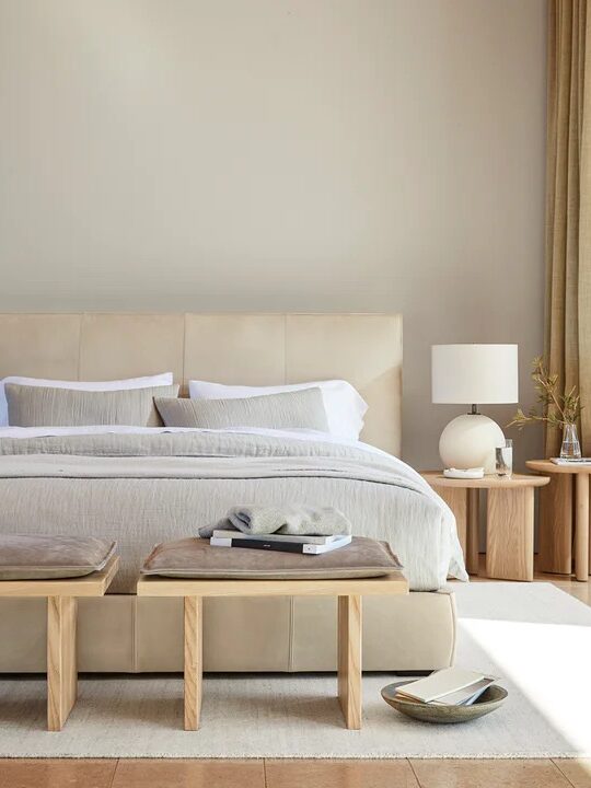 Minimalist bedroom with a beige bed, two wooden benches at the foot, two side tables with lamps, and sheer curtains.