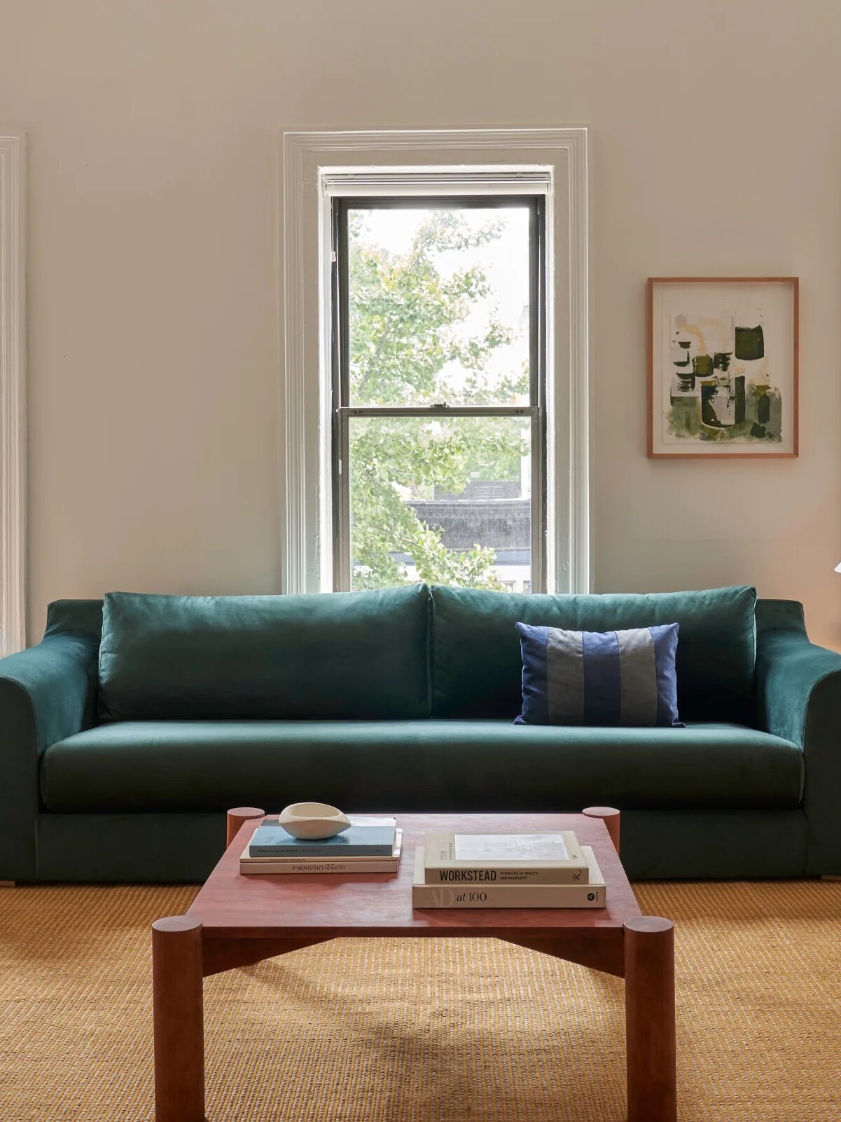 A living room with a green sofa, three windows, a wooden coffee table with books, a lamp on the floor, and a rug covering the wooden floor.