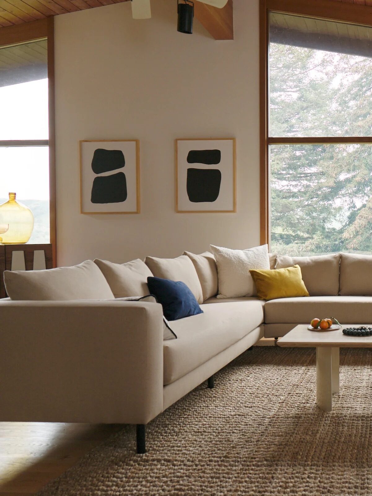 A cozy living room with a beige sectional sofa, a coffee table, and a wood-burning stove. Large windows show a view of trees. Artwork and a potted plant add decor.