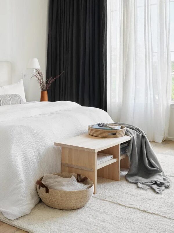 A bright bedroom with a neatly made bed featuring white linens, a small wooden table, a basket, and a gray throw. Large windows with white and black curtains overlook greenery.