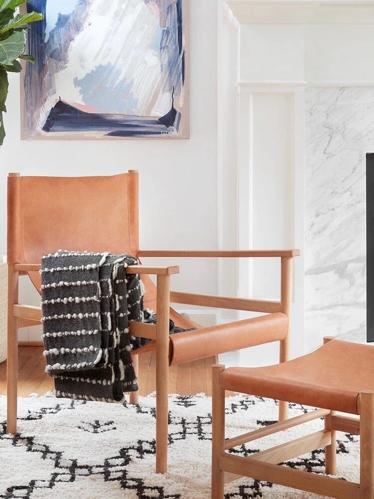 A cozy living room corner with a wooden chair, leather ottoman, and a black-and-white blanket. A painting, plant, and fireplace complete the setting on a patterned rug.