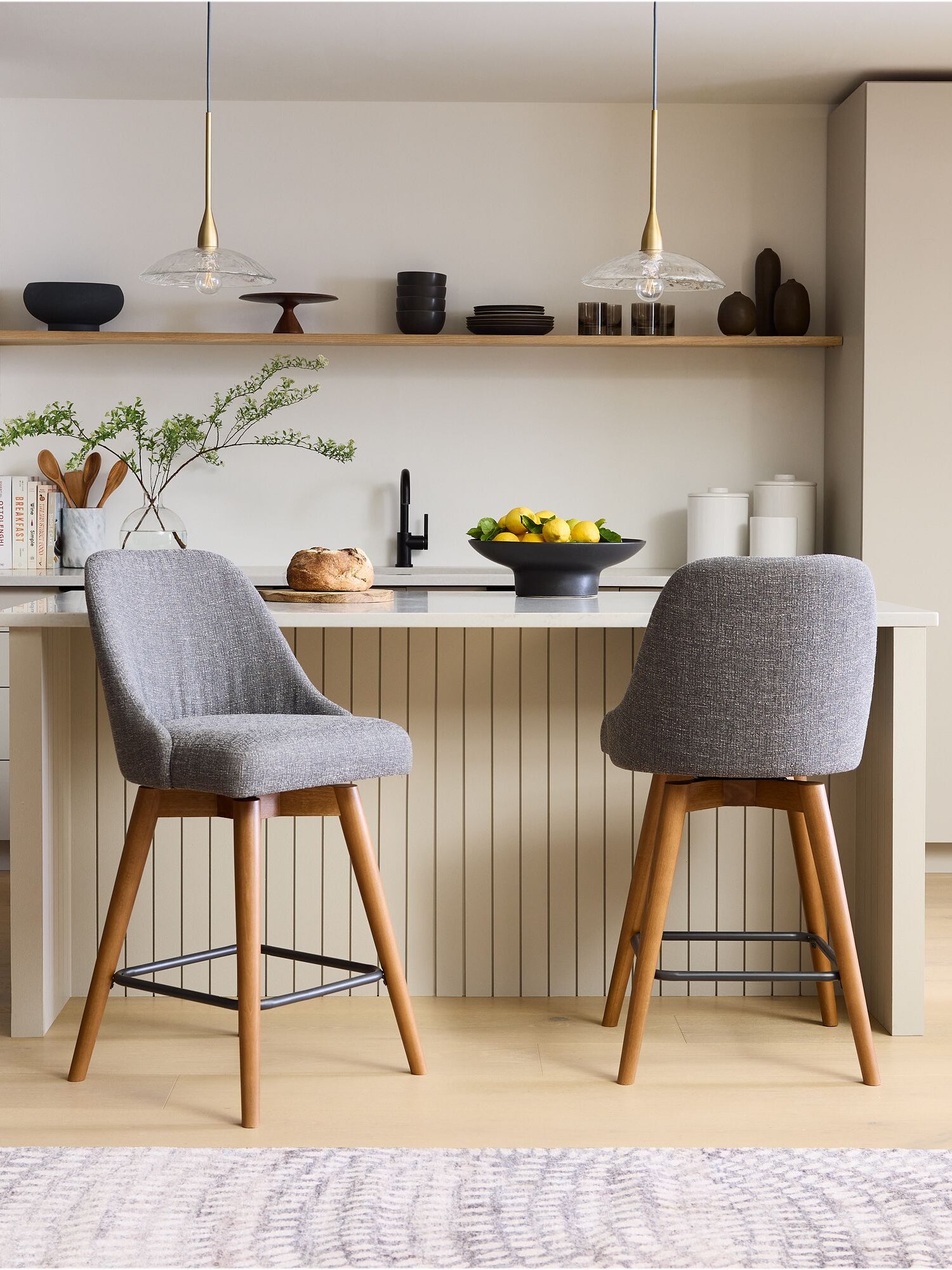 Modern kitchen with two grey upholstered barstools at a breakfast bar, pendant lights, and decorative bowls and plants on shelves.