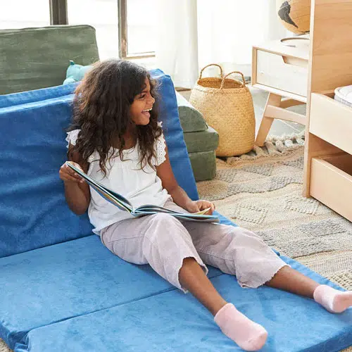 A child sits on a blue cushioned mat, smiling and holding an open book. There is a woven basket and a bookshelf in the background.