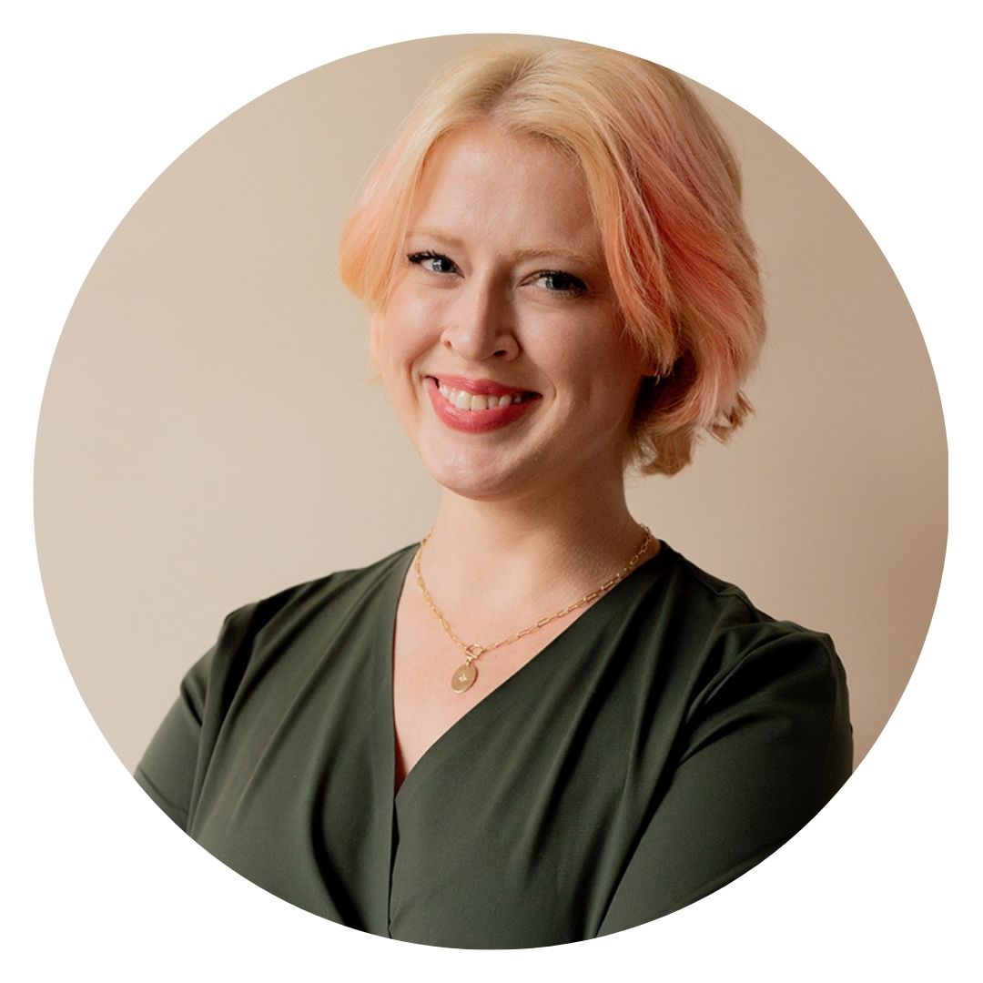 This headshot of Emily McGowan shows she has short, light hair wearing a green blouse smiles while standing with arms crossed against a neutral background.