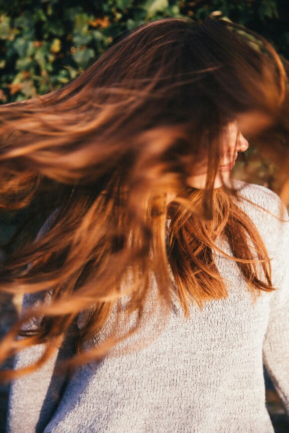 A person with long, reddish-brown hair in motion, wearing a light sweater, is standing outdoors.