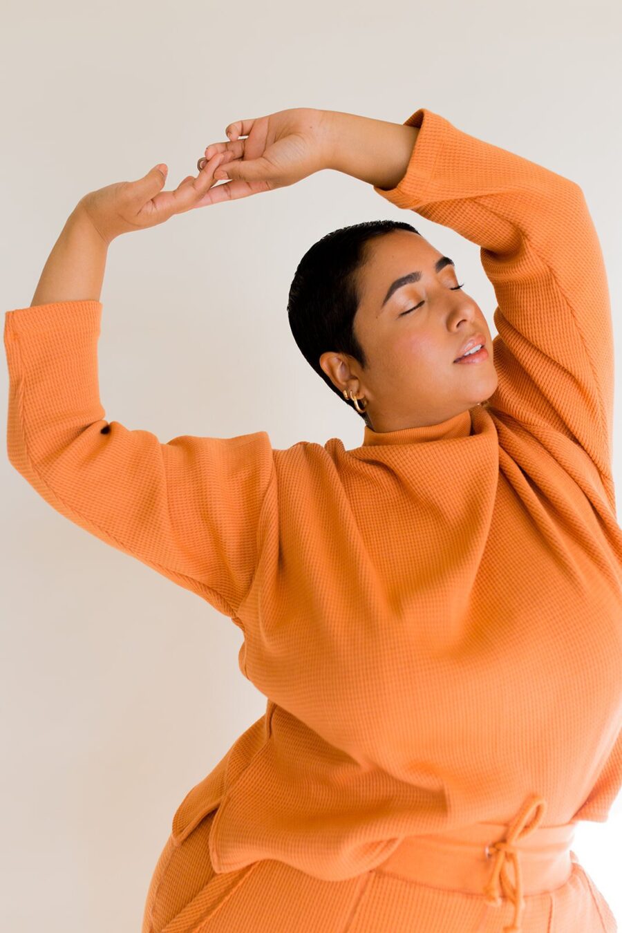 A person in an orange outfit poses gracefully with eyes closed and arms raised against a plain background.