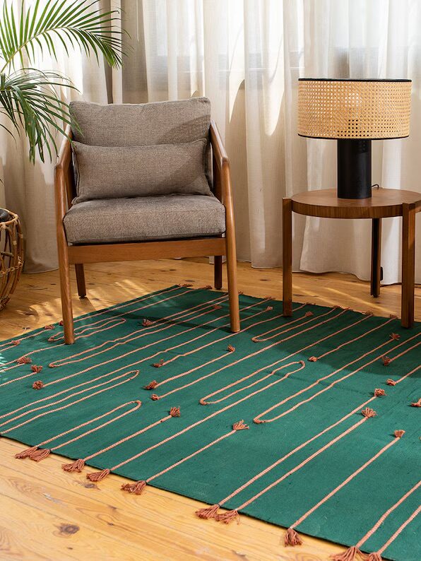 A cozy room with a gray armchair, a small wooden side table with a lamp, and a green patterned rug on a wooden floor. A plant is in the corner. Sunlight filters through sheer curtains.