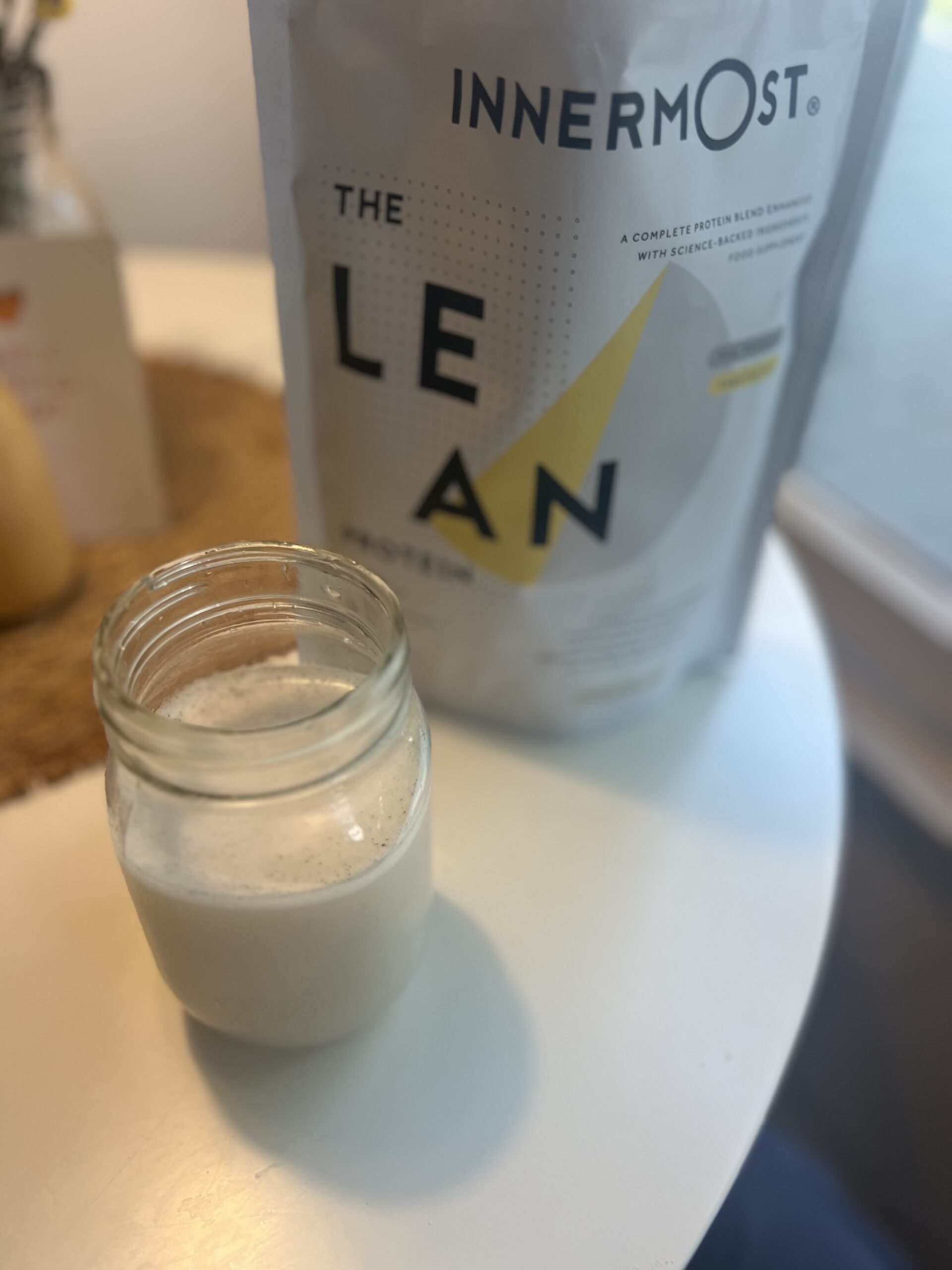 A jar filled with a creamy white liquid sits next to a package labeled "THE LEAN PROTEIN" on a table, with soft lighting from a nearby window.