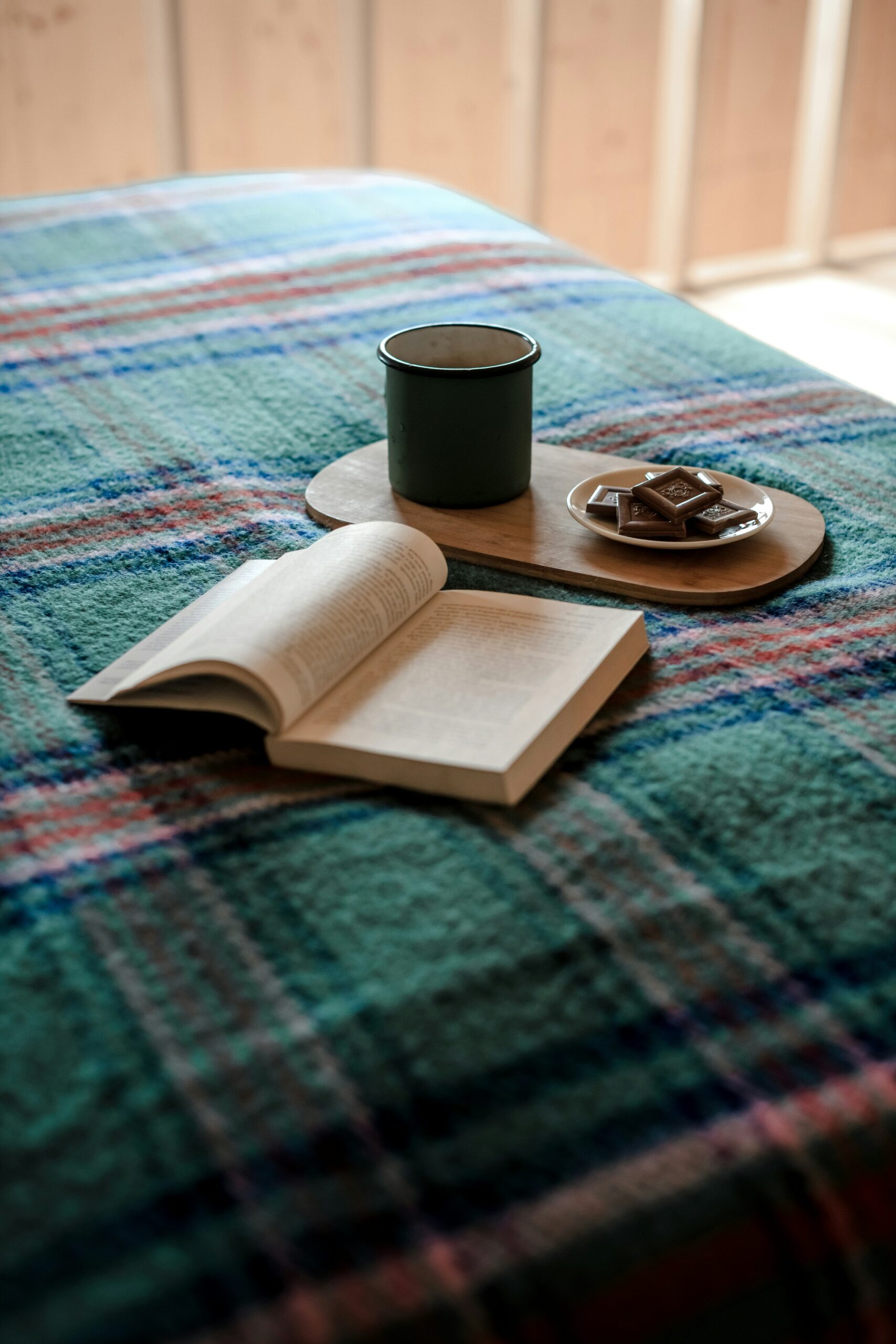 Open book on a plaid blanket with a green mug on a wooden tray and a plate of chocolates.