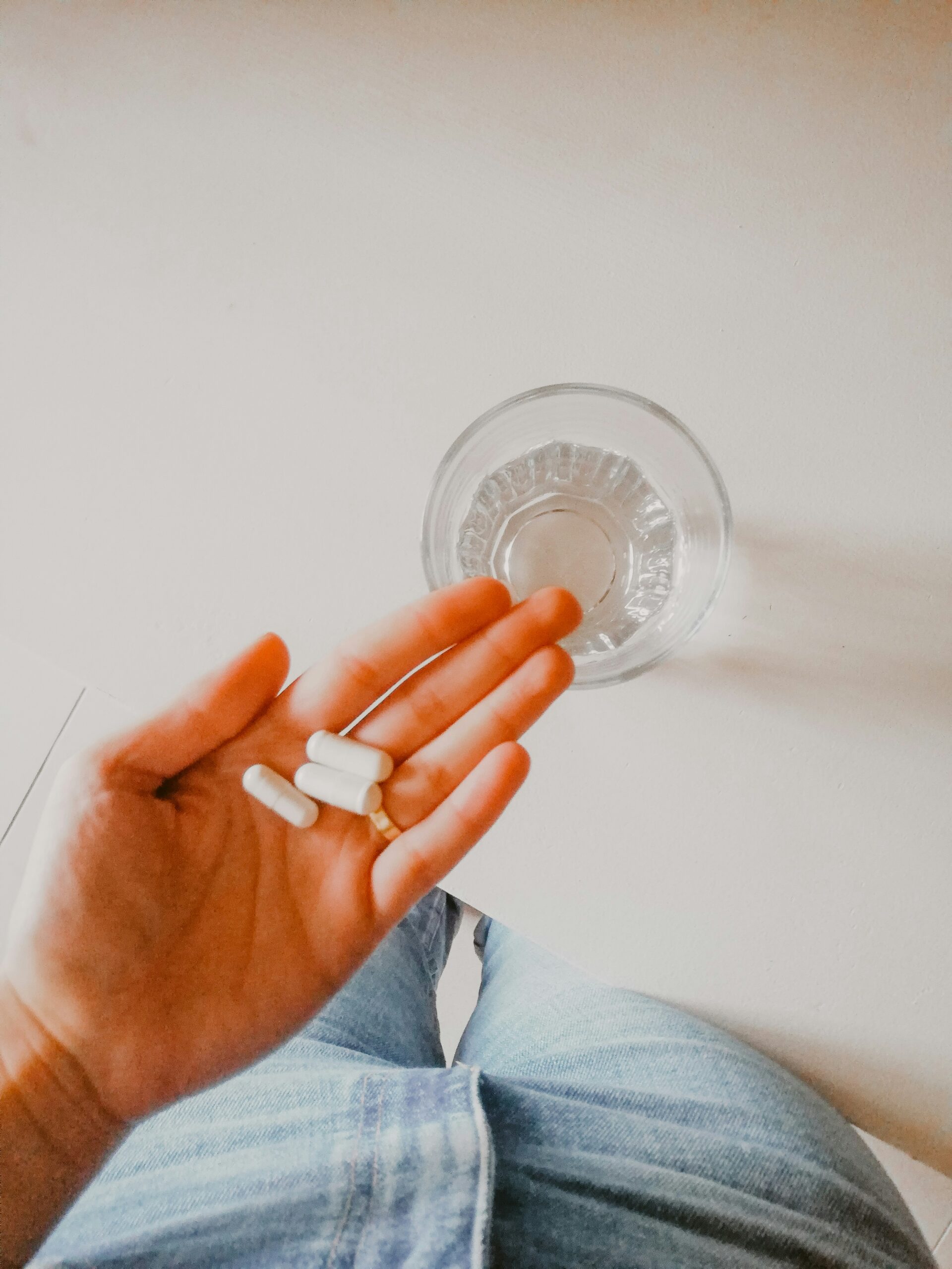 A hand holds three white capsules above a glass of water on a light surface.