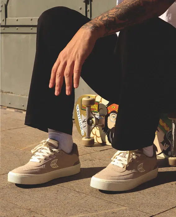 Person wearing beige sneakers and black pants, sitting on a skateboard with graphic designs.