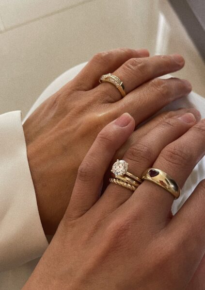 Two hands with gold rings. One hand has multiple rings, including one with a large stone, and a ring with a red stone. The other hand has a simpler band with small stones.