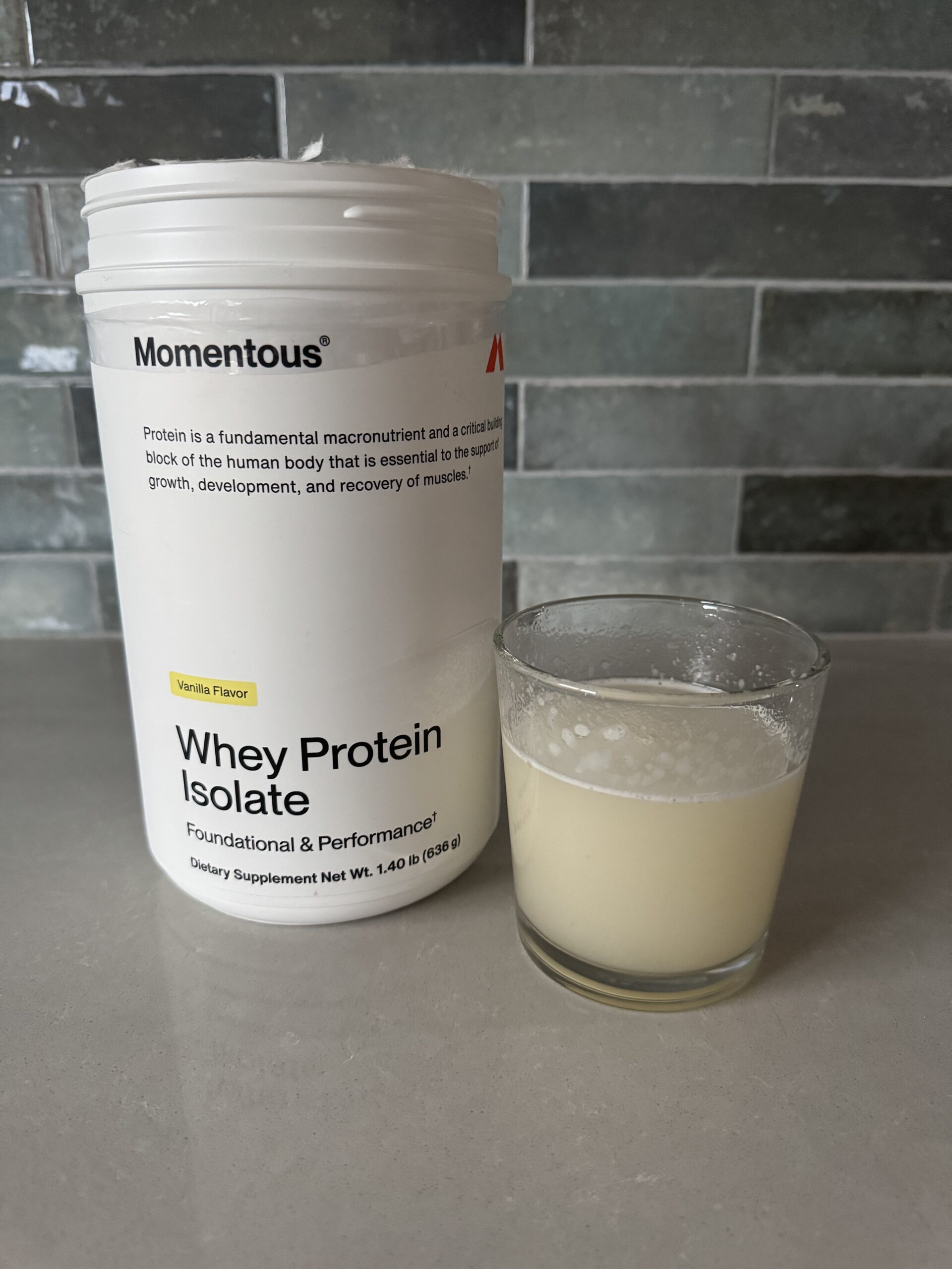 Container of vanilla whey protein isolate next to a glass filled with a light-colored liquid on a gray countertop.