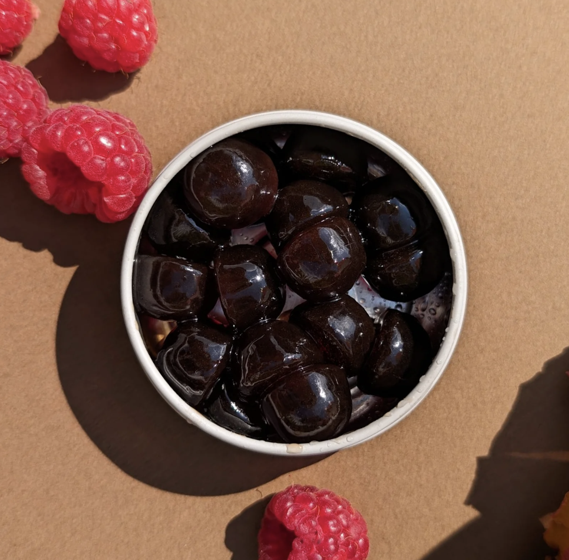 A bowl of shiny black tapioca pearls on a brown surface, surrounded by a few red raspberries.