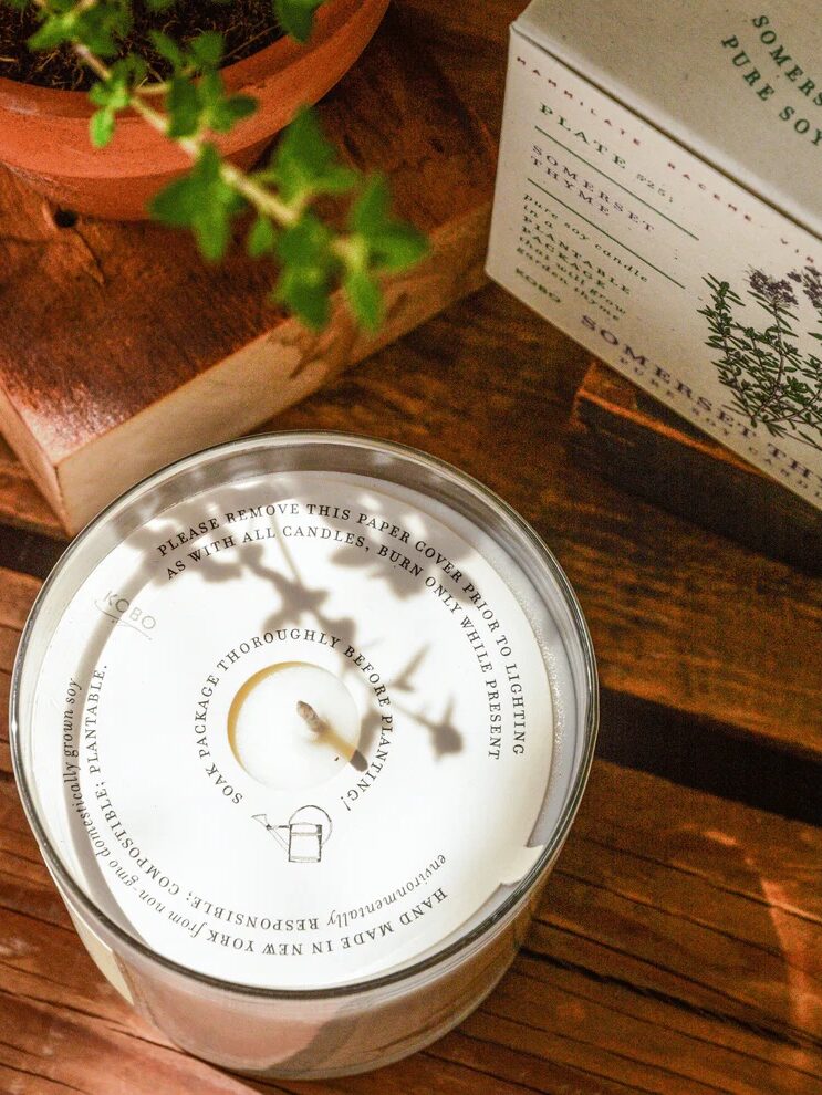 A soy candle labeled "Somerset Thyme" sits on a wooden table next to a potted plant. The candle has a white paper cover with text instructions.