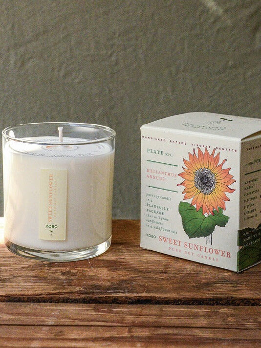 A glass jar candle labeled "Sunflower" is placed beside its decorative box on a wooden surface, with a grayish backdrop.