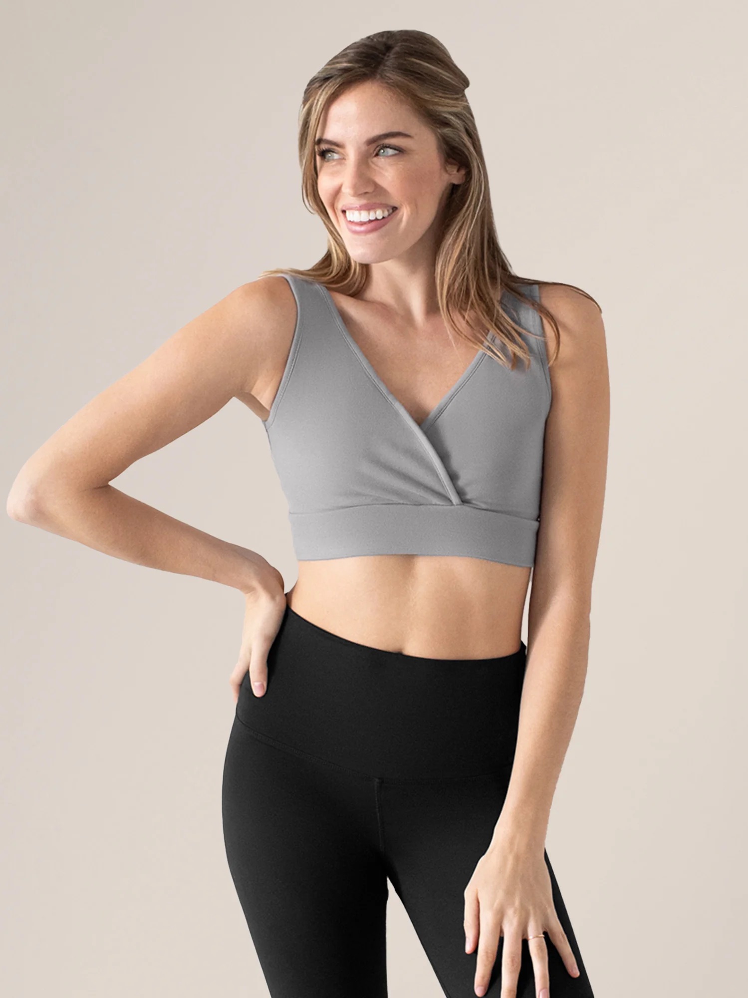 A woman wearing a gray sleeveless crop top and black leggings smiles, standing with one hand on her hip against a plain background.