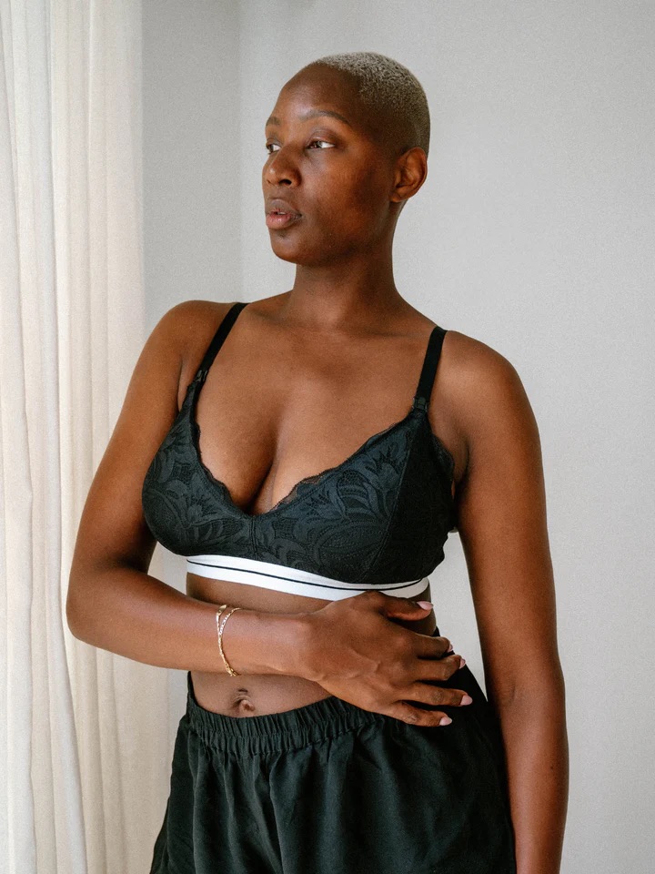 A person with short hair wearing a black bralette and black pants stands near a white curtain, looking to the side.