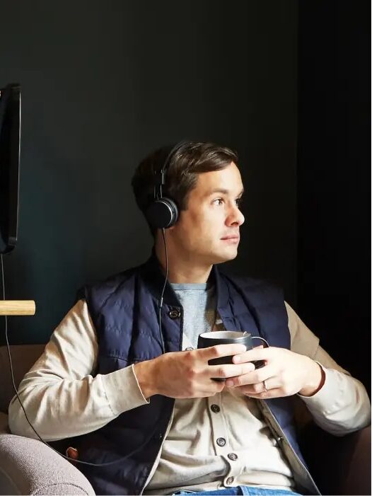 A person wearing headphones and holding a cup sits on a chair in a dimly lit room with dark walls and modern decor.