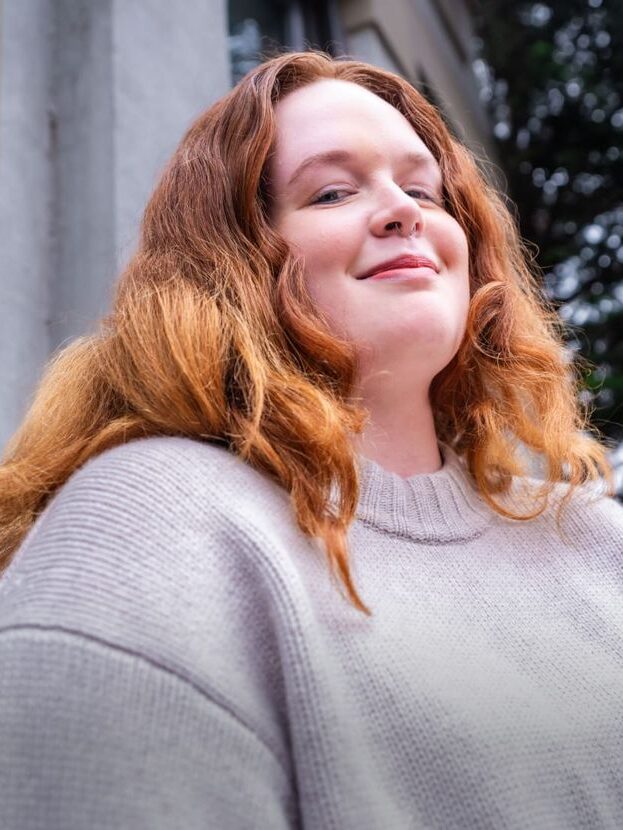 Person with long red hair wearing a gray sweater stands outside, smiling slightly. Trees and a building are in the background.