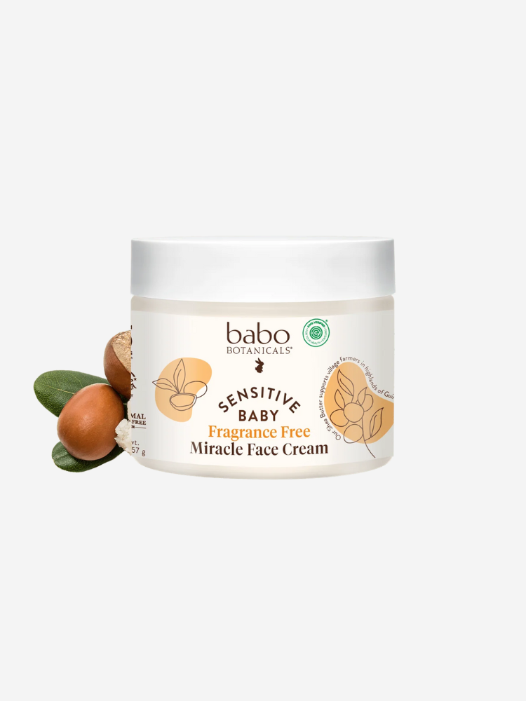 A jar of Babo Botanicals Sensitive Baby Fragrance Free Miracle Face Cream is displayed with illustrated plant elements on the label and natural ingredients beside it.