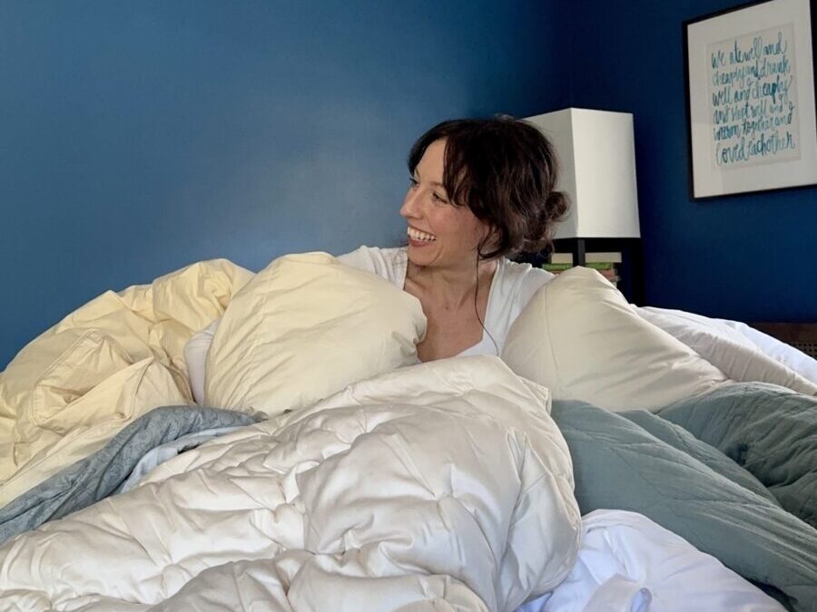 A woman smiles while sitting on a bed with an organic mattress, surrounded by multiple pillows and blankets in a room with blue walls and framed artwork.