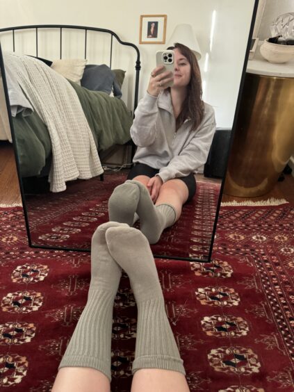 Sitting on a red-patterned rug in front of a mirror, a person wearing a gray sweater and knee-high organic socks takes a selfie with their smartphone. A cozy bed and framed picture add charm to the background.