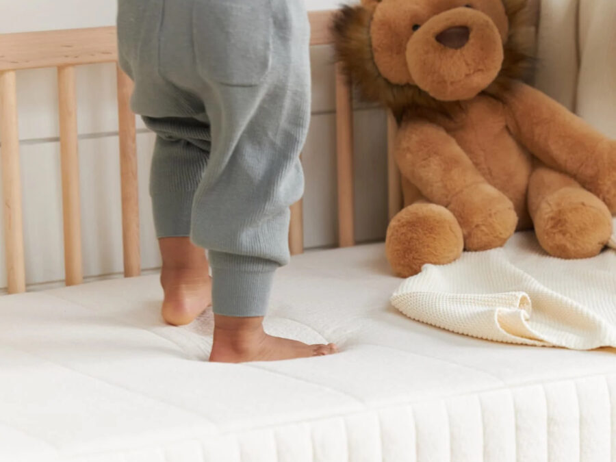 A toddler wearing gray pants stands on an organic mattress next to a plush lion toy.