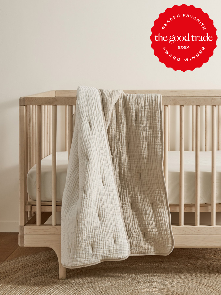 A wooden crib with a cream-colored quilt draped over the side. A red award badge in the top right corner reads "Reader Favorite The Good Trade 2024 Award Winner.