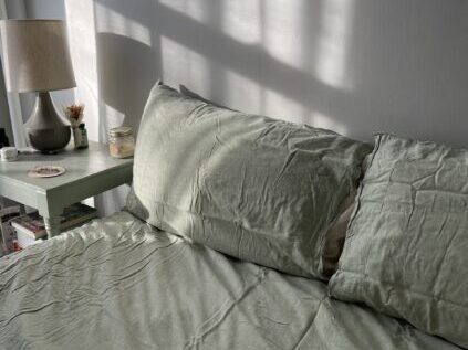 A bed with green sheets and pillows is illuminated by sunlight through a window. A bedside table holds a lamp and small items.