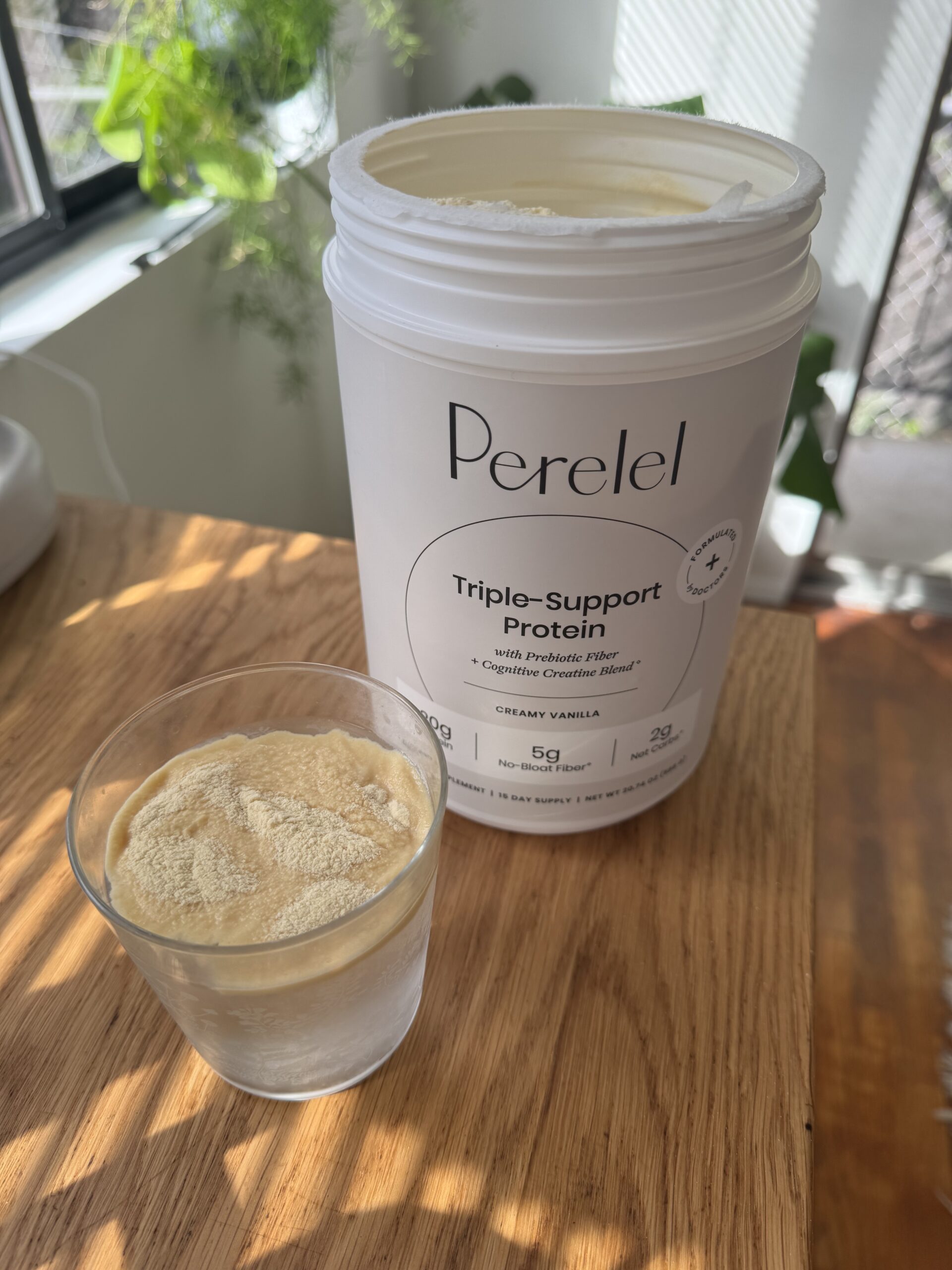 Container of Perelel Triple-Support Protein with Prebiotic Fiber on a wooden table, open with a scoop inside. A glass next to it contains a portion of the protein powder.