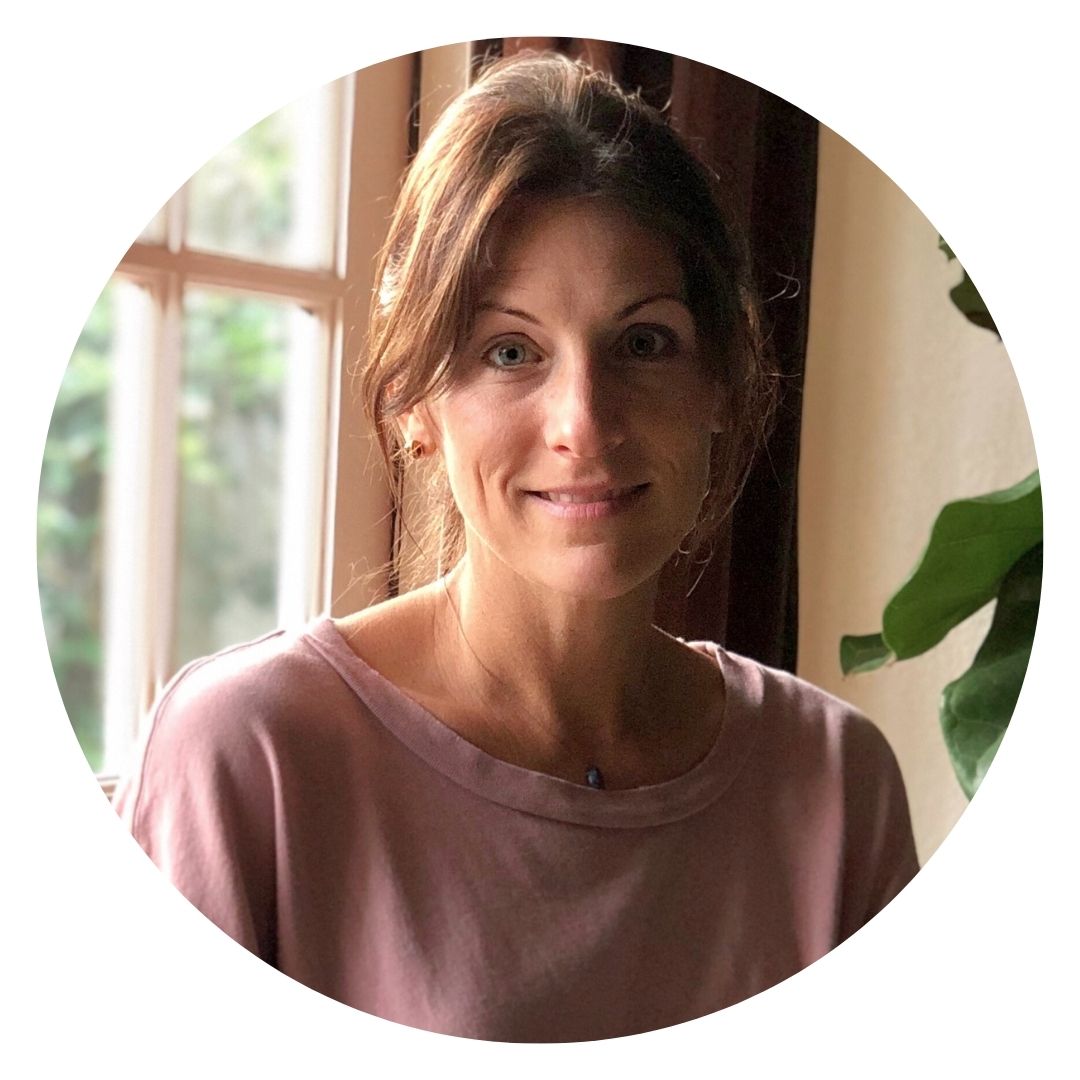 Randi Donahue is a woman with brown hair in a pink top sits near a window, reflecting on her latest mattress review, as a plant thrives in the background.