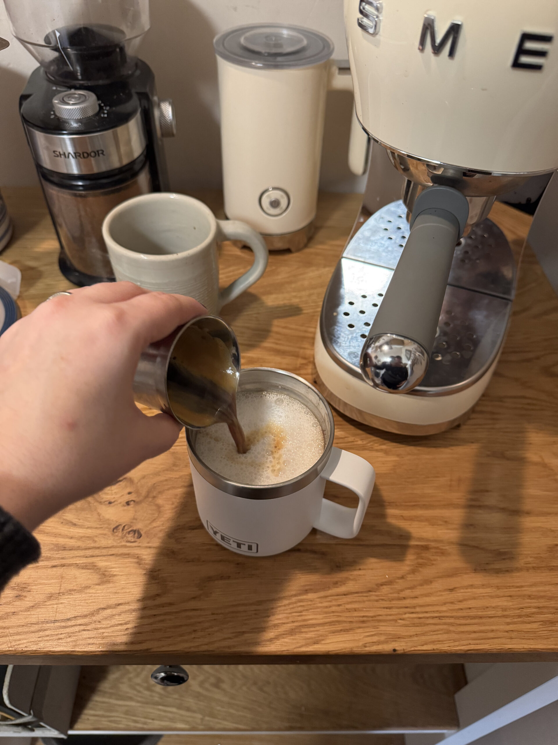 A person pours liquid from a metal cup into a mug filled with foamy milk on a wooden countertop next to a coffee maker and a mug.